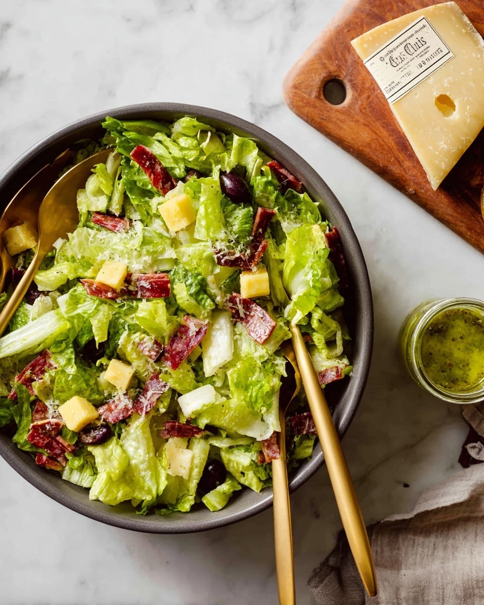 A close-up of a mixed salad in a dark bowl with chopped green lettuce as the base layer, topped with small cubes of yellow cheese, thin red slices of cured meat, dark purple olive halves, and small pieces of tomato and celery spread evenly throughout. Two golden serving spoons rest inside the bowl on top of the salad. Next to the bowl, there is a wooden board holding a wedge of pale yellow cheese with a label, and a small glass jar with green dressing. The whole scene is set on a white marbled surface. Photo taken with an iphone --ar 4:5 --v 7