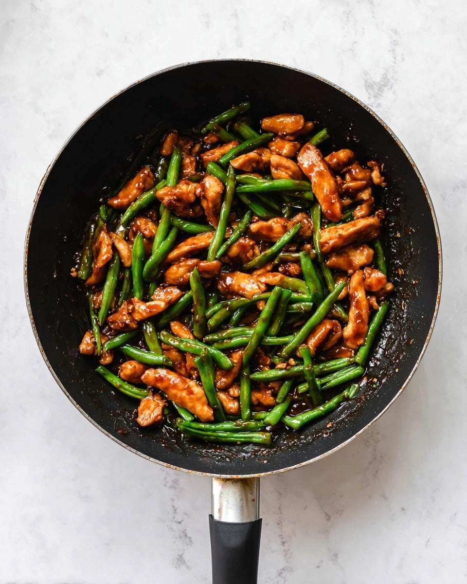 A black frying pan filled with a stir-fry dish consisting of glossy orange-brown chicken pieces and bright green string beans, both coated in a shiny dark soy sauce. The chicken pieces are irregularly shaped and scattered evenly throughout the pan, while the slender green beans lay mostly on top and intertwined with the chicken. The pan rests on a white marbled textured surface, and its handle extends downward from the frame's center. The image is bright and clear, emphasizing the vibrant colors and textures of the cooked ingredients. photo taken with an iphone --ar 4:5 --v 7