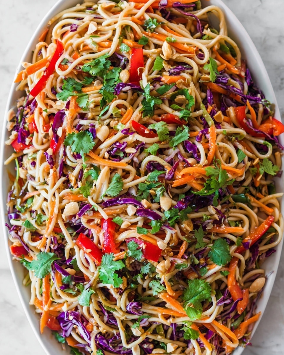 The image shows a colorful noodle salad with three main visible layers: the base layer is light beige noodles twisted and spread around, mixed throughout with thin strips of bright orange carrots and deep purple cabbage giving a crunchy texture. Scattered on top are small pieces of red bell pepper adding a fresh pop of color, alongside many fresh green cilantro leaves spread evenly for a leafy contrast. There are also small chopped nuts sprinkled throughout, adding a rough texture among the smooth noodles and crunchy veggies. The dish rests on a white plate over a white marbled surface. photo taken with an iphone --ar 4:5 --v 7