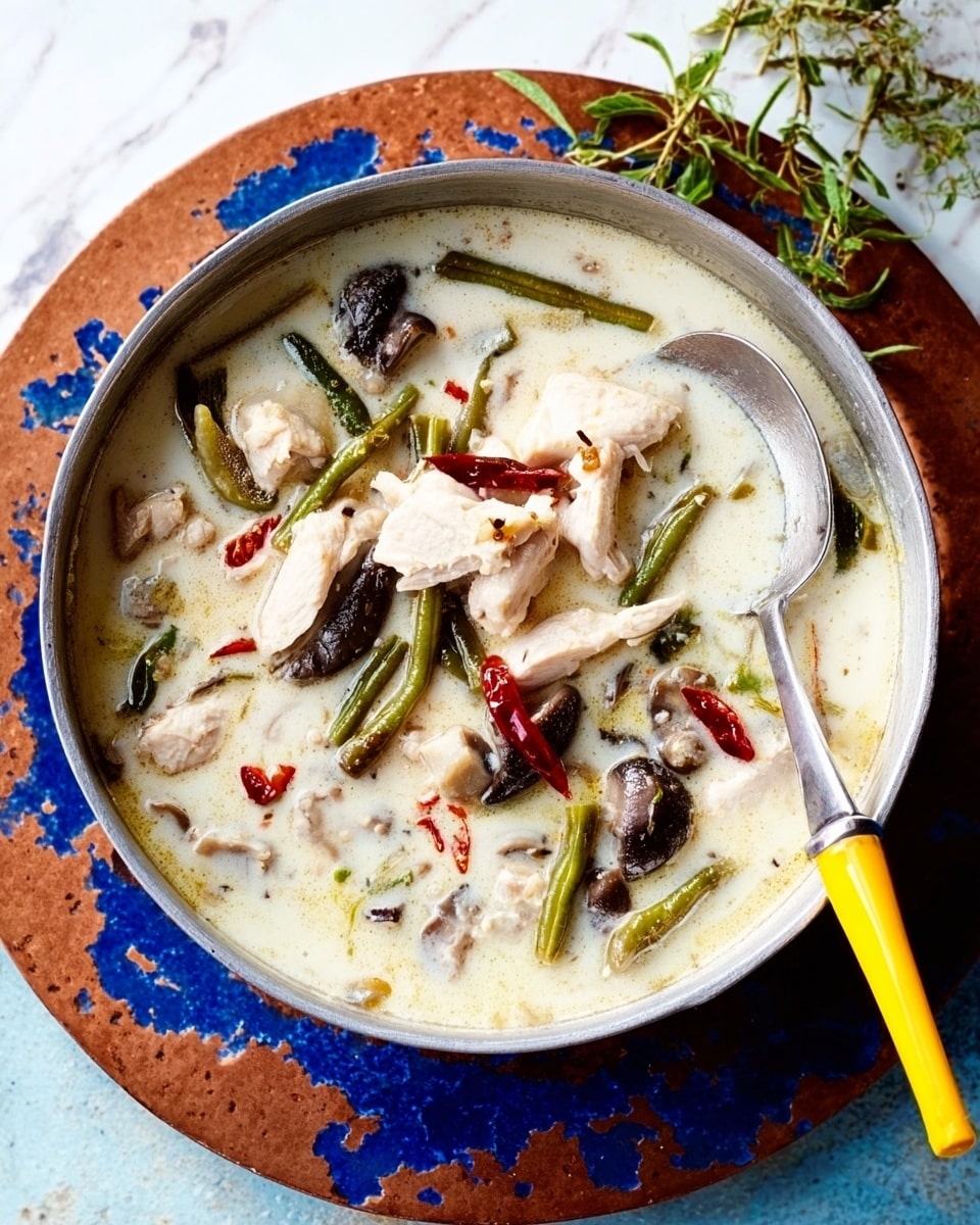 The image shows a round silver pot filled with a creamy white soup that has many pieces of white chicken, dark brown mushroom slices, and long green beans scattered throughout. There are small red chili pieces on top which add a pop of color. A silver ladle with a yellow handle rests inside the pot on the right side. The pot is placed on a round rusty brown tray with blue spots, all set on a white marbled surface. A small bunch of green herbs is seen on the top right outside the pot. photo taken with an iphone --ar 4:5 --v 7