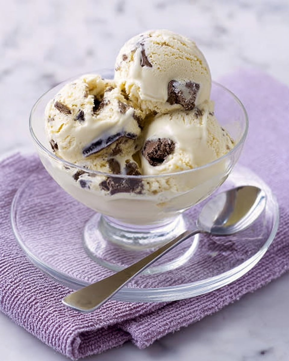 A clear glass bowl filled with three scoops of creamy vanilla ice cream with dark chocolate chunks mixed inside. The bowl sits on a clear glass plate, which is placed on a light purple cloth with a woven pattern. A shiny silver spoon rests on the right side of the plate. The scene is set on a white marbled surface. Photo taken with an iphone --ar 4:5 --v 7