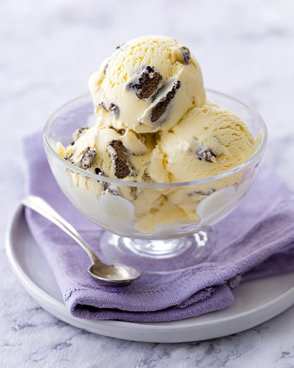 The image shows three round scoops of light yellow ice cream with visible chunks of dark chocolate or cookie pieces mixed inside, all placed neatly in a clear glass bowl. The bowl is set on a white plate with a matching clear texture, resting on a folded lavender cloth. Next to the plate lies a small silver spoon. The background is a white marbled surface that adds a soft contrast to the creamy ice cream colors. photo taken with an iphone --ar 4:5 --v 7