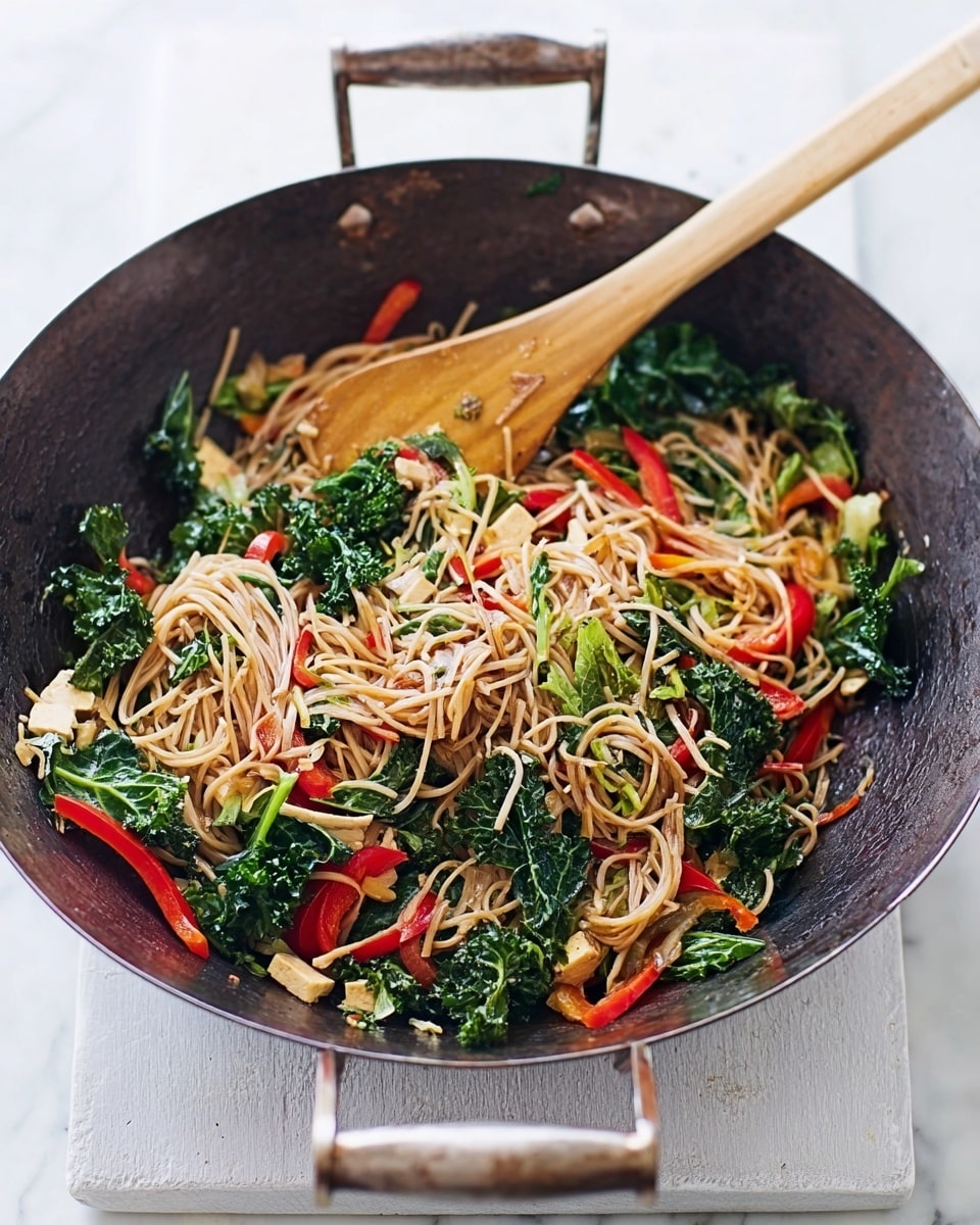 A large dark metal wok filled with cooked noodles mixed with various colorful vegetables sits on a white marbled surface. The dish has several layers: at the bottom are light brown noodles with a smooth texture, mixed with bright green kale leaves that are slightly wilted and curly, red bell pepper slices adding a vibrant splash, and small chunks of light beige tofu scattered throughout. A wooden spatula rests inside the wok, stirring the ingredients, with its light wooden color contrasting against the dark wok and colorful food around it. The scene is bright and clean, showing the rustic wok on a white marbled background. photo taken with an iphone --ar 4:5 --v 7