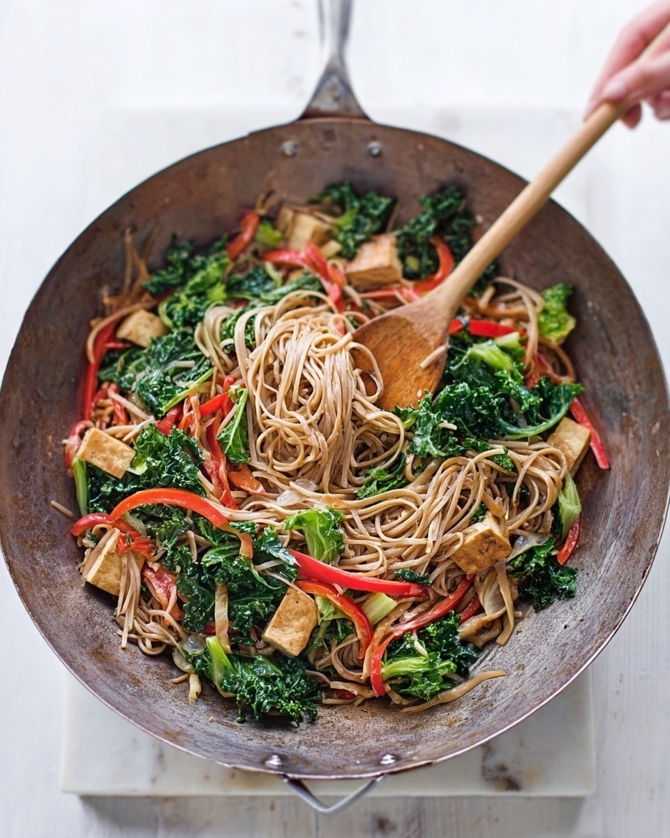 A round metal wok filled with light brown noodles mixed with bright green kale leaves, red bell pepper strips, and light golden brown pieces of tofu, all stirred with a wooden spatula held by a woman's hand. The dish shows a mix of soft, leafy texture and smooth noodles contrasted by the firm tofu chunks. The wok rests on a white marble textured surface, with a clean, bright background enhancing the colors of the food. photo taken with an iphone --ar 4:5 --v 7
