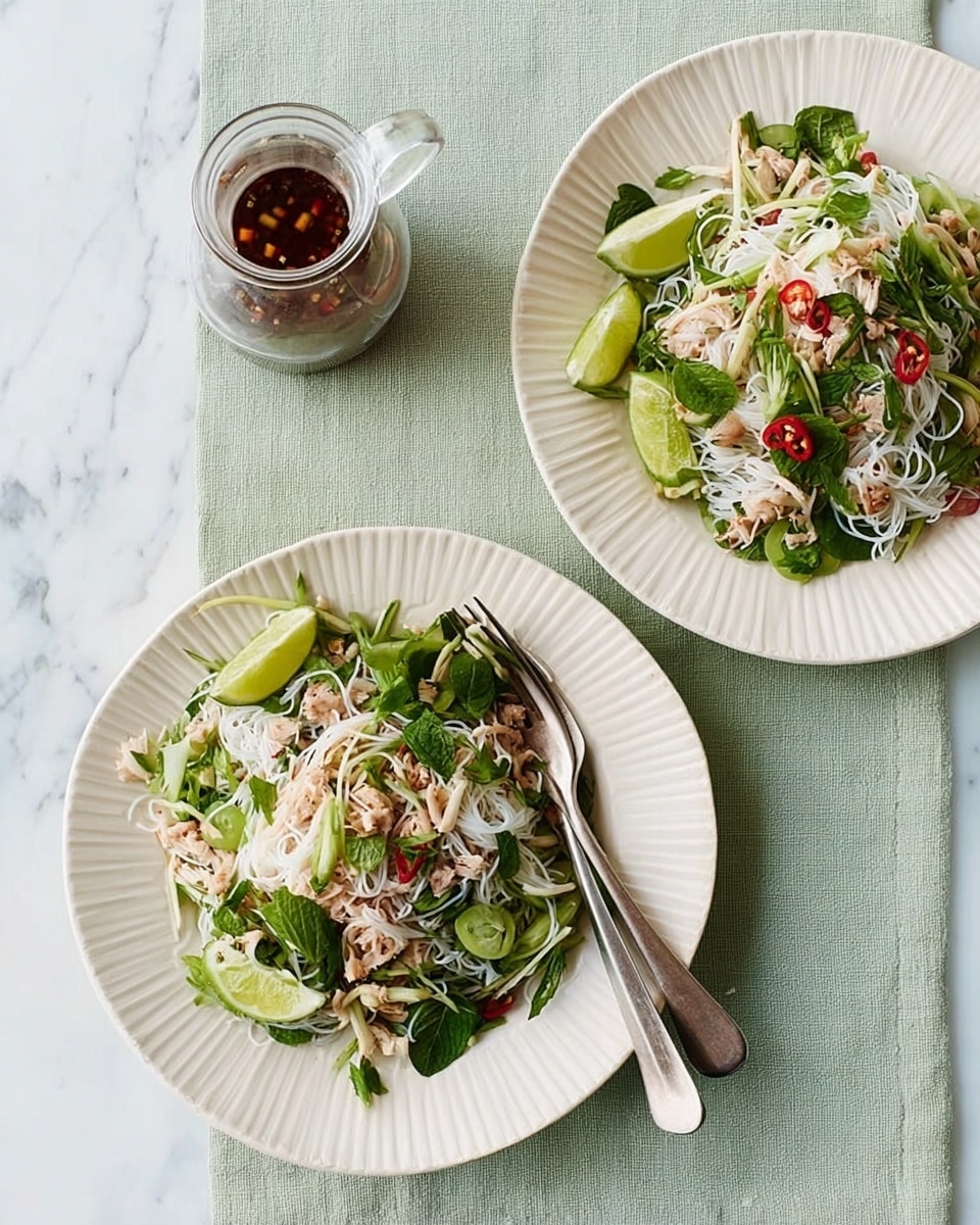 Two white round plates with ridged edges sit on a pale green cloth over a white marbled surface. Each plate holds a fresh salad with a base layer of white rice noodles mixed with green herbs and thinly sliced vegetables. The salad includes small pieces of light brown chicken or crab meat, red chili slices, and green lime wedges placed on the side. Near one plate, two silver forks rest together. Above the plates, there is a small clear glass bottle with a handle, holding dark brown sauce. photo taken with an iphone --ar 4:5 --v 7