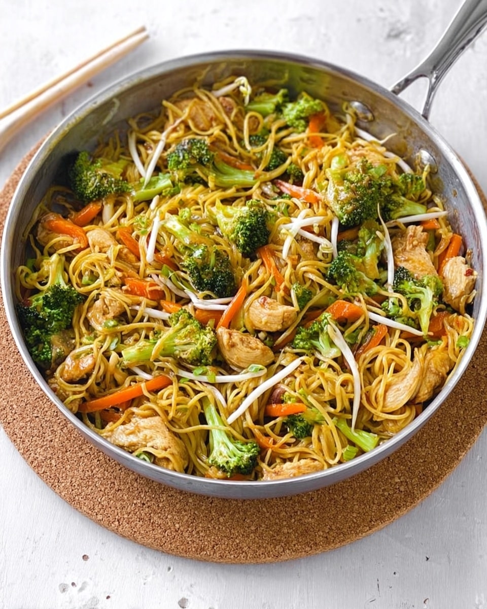 A large silver pan filled with stir-fried noodles, with a mix of bright green broccoli florets, orange carrot sticks, and small pieces of light brown cooked chicken scattered throughout. There are also white bean sprouts layered on top, adding a fresh look. The noodles are golden-yellow and mixed evenly with the vegetables and chicken. The pan sits on a round cork mat, all placed on a white marbled textured surface. photo taken with an iphone --ar 4:5 --v 7