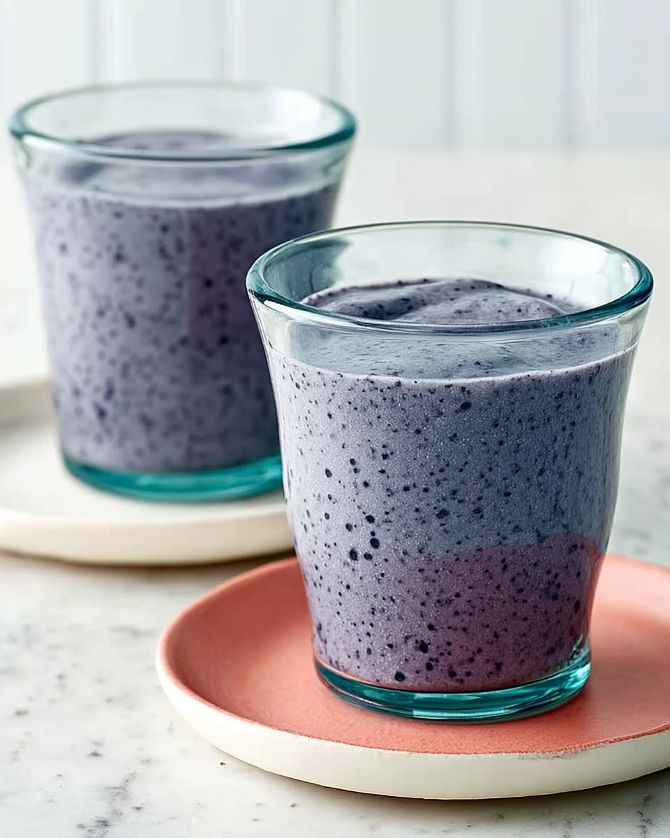 The image shows two clear glass cups filled with a thick, bluish-purple smoothie that has small dark specks throughout, indicating blended berries or seeds. Each cup sits on a small white plate with a coral-colored surface. The background and surface are a white marbled texture, providing a clean and bright setting. The overall look is fresh and inviting, with the creamy texture of the smoothie clearly visible in the glasses. Photo taken with an iphone --ar 4:5 --v 7