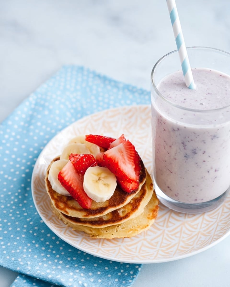 The image shows a close-up of a small stack of golden-brown pancakes topped with slices of banana and fresh red strawberry pieces arranged neatly on a white plate with a simple geometric pattern. To the right, there is a clear glass filled with a creamy light purple smoothie that has visible tiny fruit specks, with a white and blue striped straw inside. The plate and glass sit on a white marbled surface with a light blue cloth that has small white dots on the left side. Photo taken with an iphone --ar 4:5 --v 7