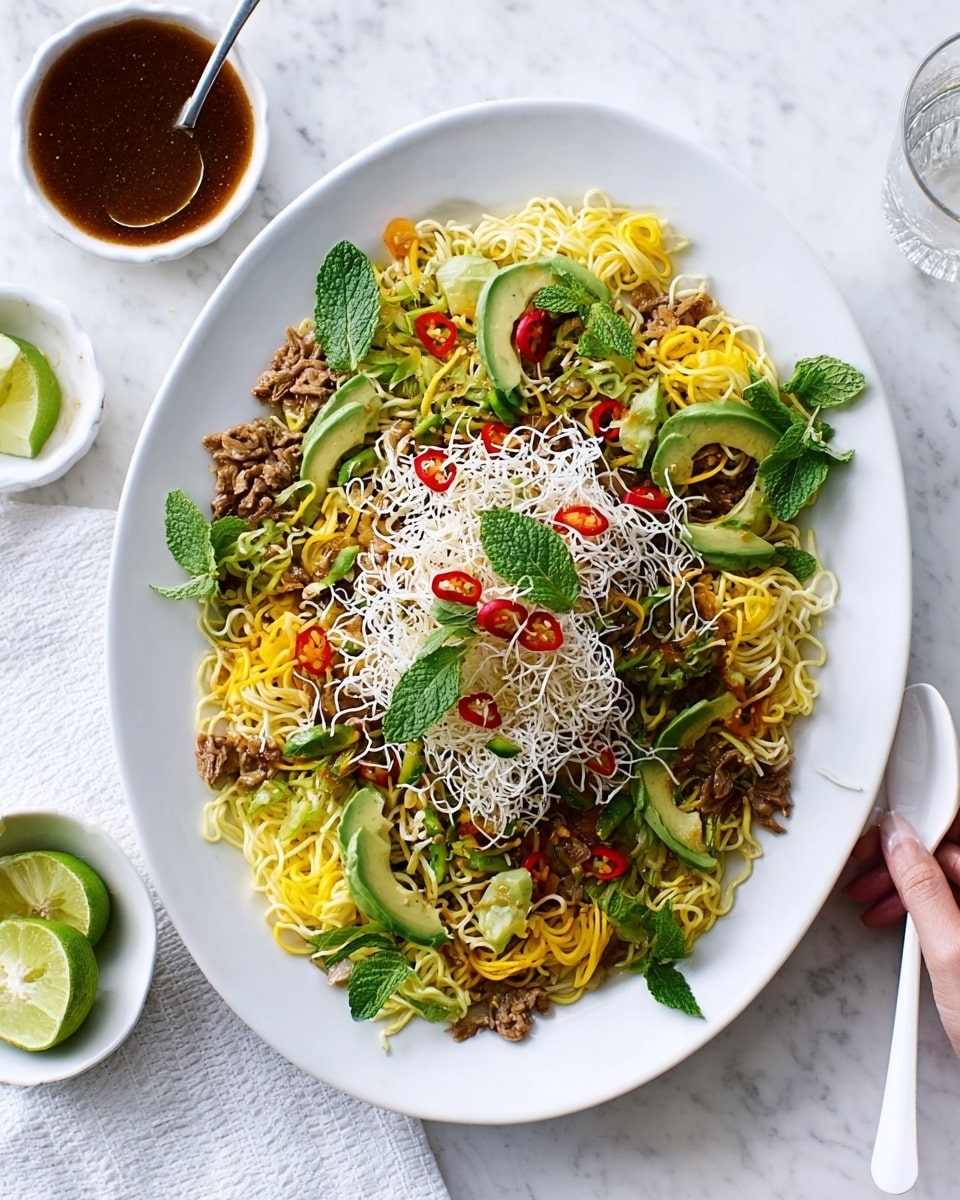 A white oval plate holds a layered noodle dish starting with a base of yellow cooked noodles spread across the plate. On top, there are slices of green avocado and thin strips of red chili, adding bright spots of color. Fresh green mint leaves are scattered over the dish, giving a fresh look. The top layer is made of crunchy white fried noodles piled in the center. Around the plate, small pieces of cooked brown meat and some yellow vegetables add texture and depth. The plate sits on a white marbled surface, with a small bowl of dark brown sauce with a spoon above it, a glass of lime on the left, and a woman's hand holding a white fork and spoon to the side. photo taken with an iphone --ar 4:5 --v 7