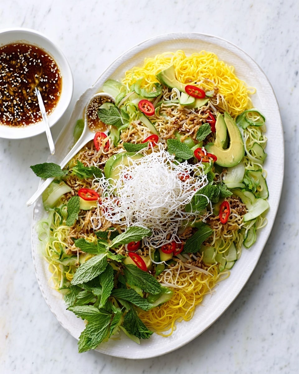 A white oval plate sits on a white marbled surface, filled with a vibrant layered dish. The bottom layer consists of bright yellow noodles spread evenly across the plate. On top of the noodles, there are slices of green avocado, scattered fresh mint leaves, red chili pieces, and thinly sliced pale green vegetables. The dish is finished with a generous mound of crispy white fried noodles piled in the center. To the side, there is a small white bowl filled with a dark brown sauce containing sesame seeds and small bits of herbs, with a white spoon resting inside it. Photo taken with an iphone --ar 4:5 --v 7