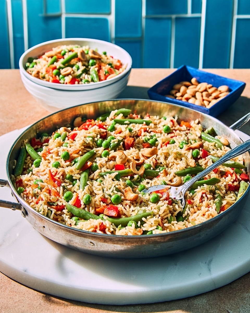 A large metal pan filled with a colorful vegetable rice mixture sits on a white round tray. The dish has layers of cooked rice mixed with bright green beans, chopped red bell peppers, green peas, and a few light brown nuts, giving it a fresh and vibrant look. A metal spoon is stuck into the pan, lifting some of the rice. Next to the pan, there is a white bowl with a similar rice and vegetable mix, placed on a white marbled surface. A blue square dish with light brown nuts sits close by. The background shows blue tiled walls. Photo taken with an iphone --ar 4:5 --v 7