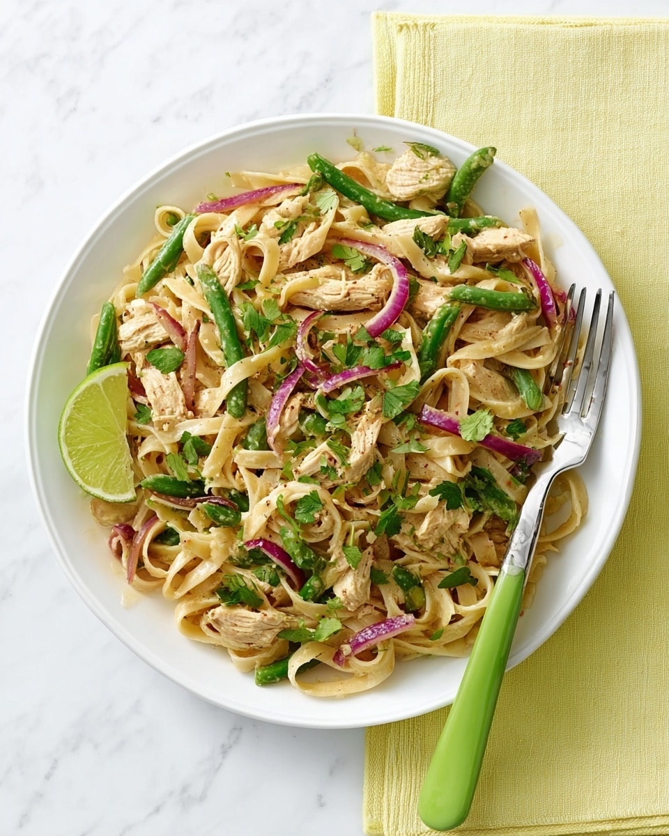 A white round plate filled with a colorful noodle dish, featuring flat light beige noodles layered with thin strips of cooked light brown chicken, small pieces of green beans, sliced red onions, and chopped bright green herbs on top. A half lime with a pale green inside rests on the edge of the plate, while a green-handled fork is placed on the right side. The plate is set on a white marbled surface with a pale yellow napkin partially under it. photo taken with an iphone --ar 4:5 --v 7