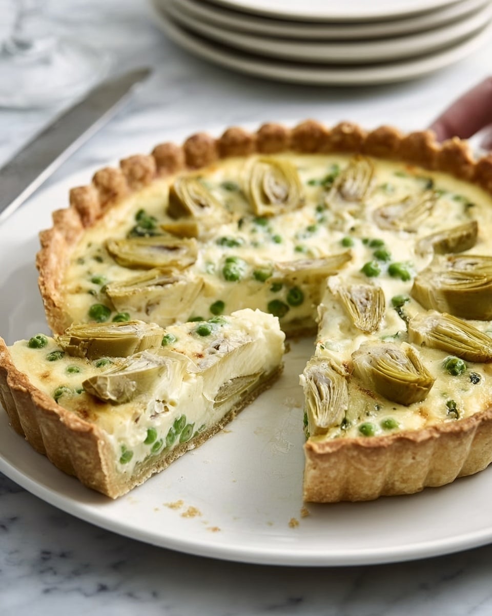 A white plate holds a savory tart with a golden, thick crust that has slightly crimped edges. The tart has a creamy, pale yellow filling dotted with green peas and topped with halved artichokes arranged evenly on the surface. One slice has been cut and slightly pulled out with a knife that rests on the plate. The background shows a white marbled surface with stacked plates blurred in the distance, and a woman's hand is just touching the edge of the tart. Photo taken with an iphone --ar 4:5 --v 7