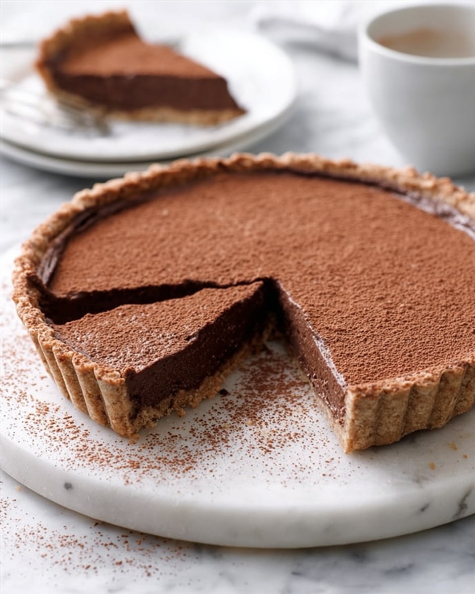 A chocolate tart with a crumbly crust and smooth, thick chocolate filling sits on a round white marble board. The tart has a dusting of cocoa powder evenly spread on top, giving it a soft brown layer above the dark chocolate inside. One slice is cut out, showing the rich, dense texture of the filling and the firm, crumbly crust edge around it. In the background, a white cup and a white plate with the removed tart slice are placed on a white marbled surface. The photo taken with an iphone --ar 4:5 --v 7