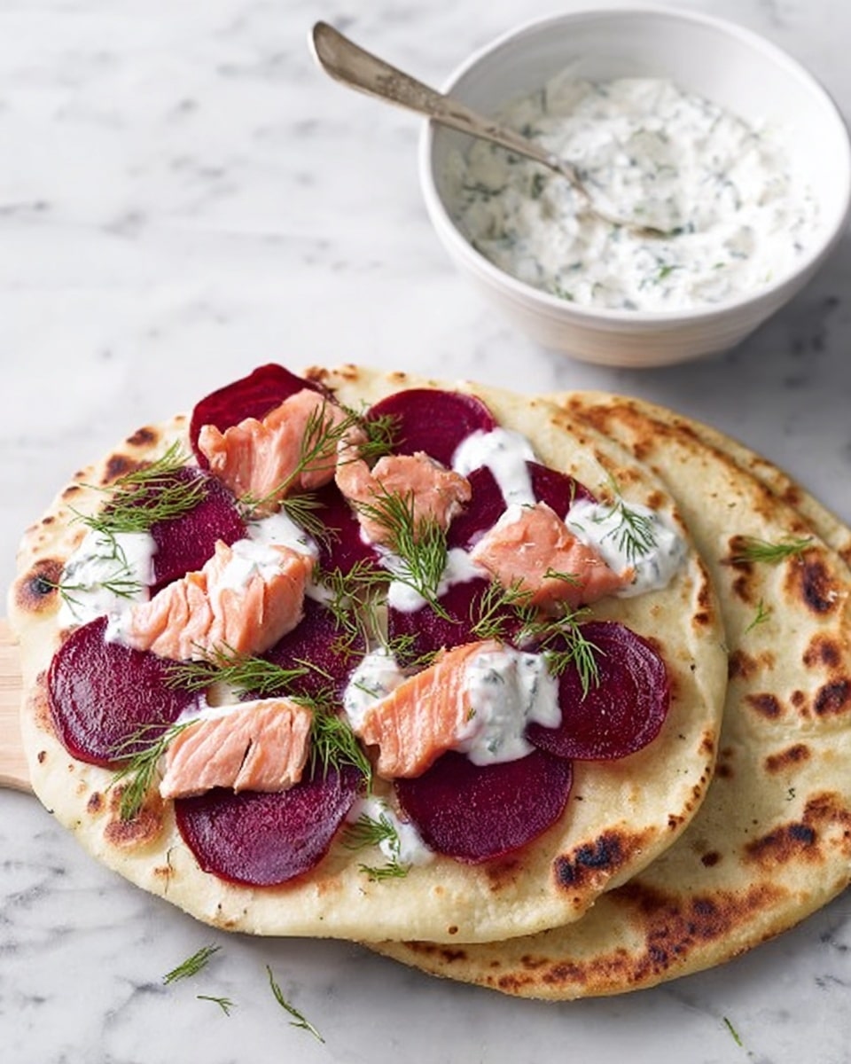 A flatbread with two visible layers: the bottom layer is a light golden-brown flatbread with some darker toasted spots; on top of it is a second flatbread spread with thin, round slices of deep red beetroot evenly placed, layered with chunks of light pink cooked salmon, and drizzled with white creamy sauce. Small green dill sprigs decorate the dish on top. Next to the flatbreads, there is a white bowl with a spoon filled with white creamy sauce containing green herbs, set on a white marbled surface. Photo taken with an iphone --ar 4:5 --v 7