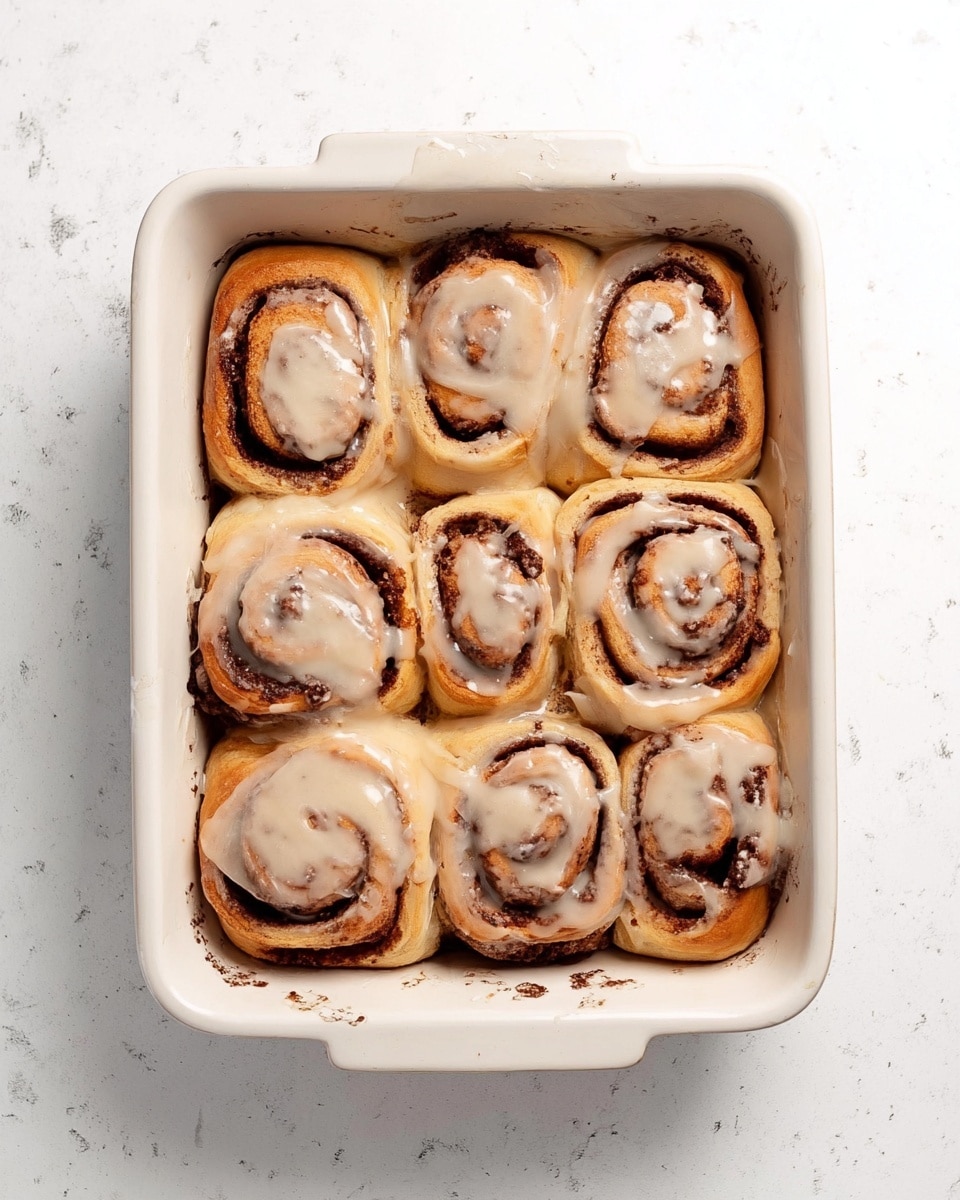 Eight cinnamon rolls are placed inside a white rectangular baking dish on a white marbled surface. Each roll is swirled with dark brown cinnamon filling and topped with a light brown creamy frosting that looks smooth and slightly melted, covering most of the rolls unevenly. The dough is golden brown and soft-looking, with some areas showing a light crust. The rolls are packed close together, filling the whole dish. photo taken with an iphone --ar 4:5 --v 7