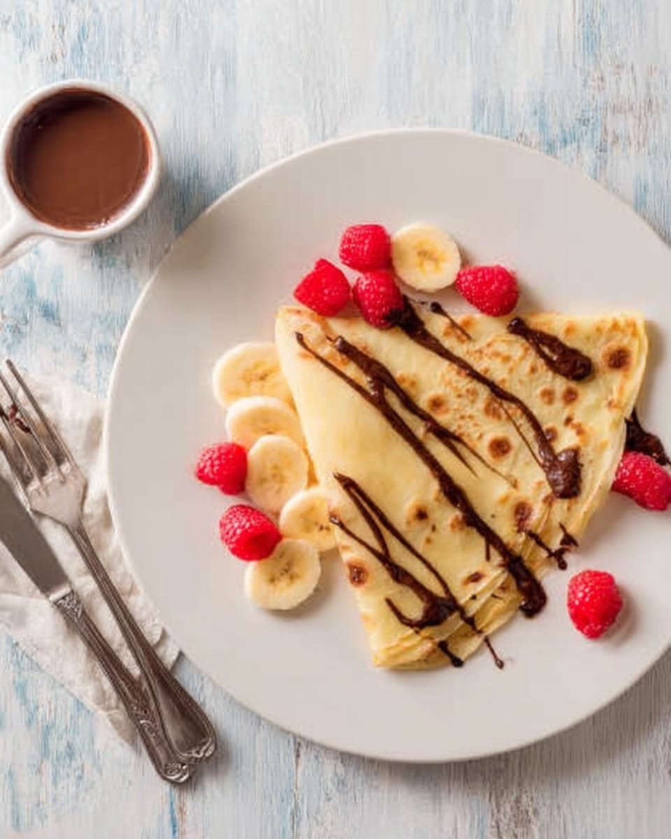 A white plate holds two folded crepes placed side by side, with the top crepe showing a light golden brown color and smooth, thin texture. On the crepes, sliced banana pieces and fresh red raspberries are scattered, some covered in a dark, glossy chocolate sauce drizzled unevenly on top. A shiny silver fork and knife rest on the right edge of the plate. Beside the plate, a small white jug with more chocolate sauce is partially visible. The whole setup is on a white marbled surface. photo taken with an iphone --ar 4:5 --v 7