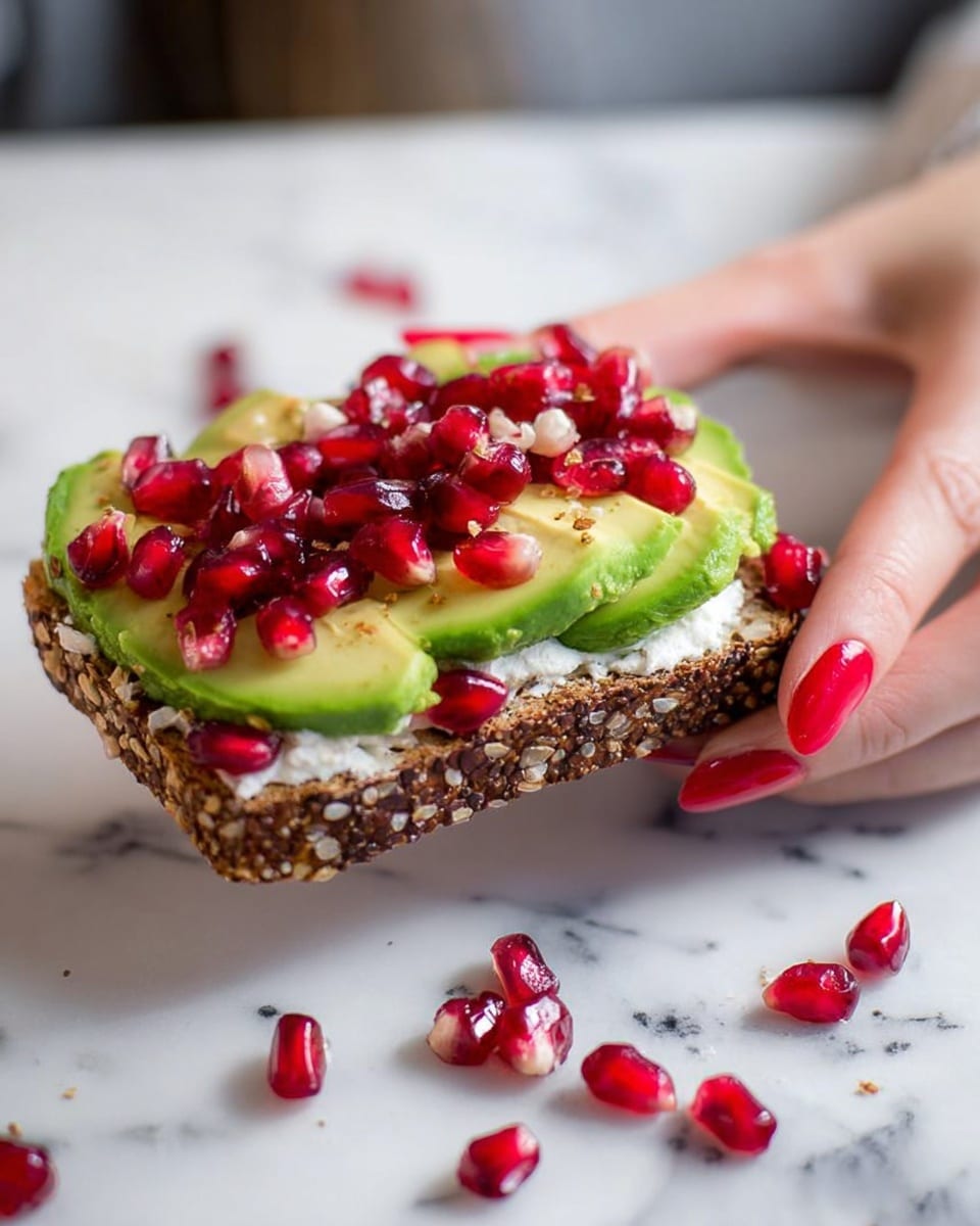 A woman's hand with red-painted nails is holding a slice of dark brown seeded bread. The bread has three layers: the bottom layer is a thick white spread, the middle layer is sliced green avocado arranged neatly, and the top layer is bright red pomegranate seeds scattered generously. Some seeds are also scattered around the bread on a white marbled surface. Photo taken with an iphone --ar 4:5 --v 7