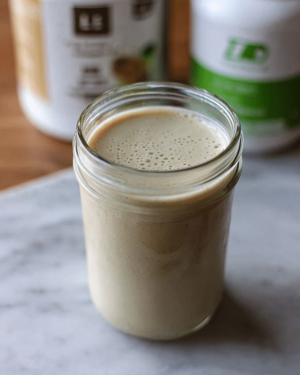 A clear glass jar filled to the top with a creamy, light beige smoothie with a smooth texture and a few small bubbles on the surface. The jar is placed on a wooden surface with blurred containers in the background, one with a green and white label and the other with a beige and green label. The focus is on the jar while the background is soft and out of focus. photo taken with an iphone --ar 4:5 --v 7