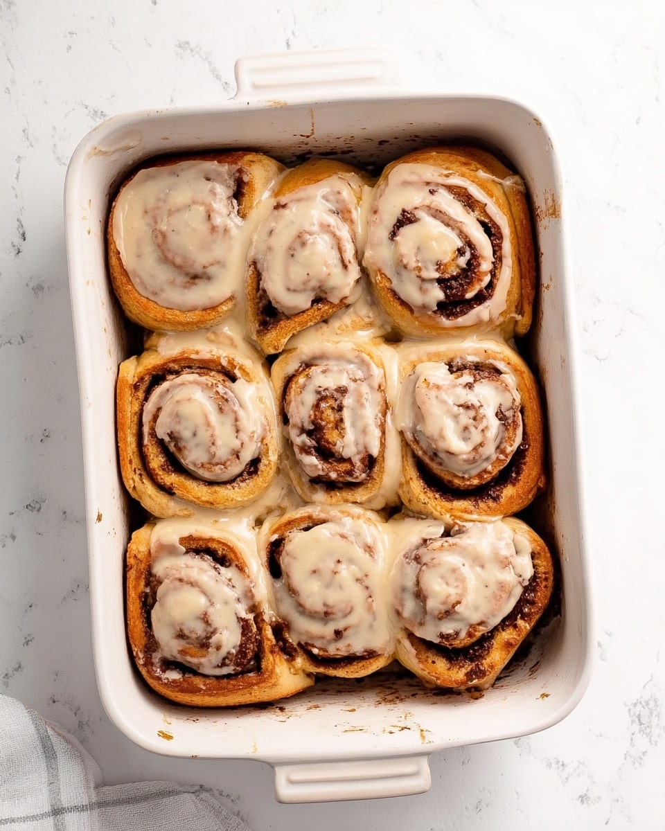 A white rectangular baking dish holds eight cinnamon rolls closely placed in two rows of four. Each roll has a golden-brown outer layer with a darker brown cinnamon swirl visible within. The rolls are topped with a smooth, light tan cream cheese frosting that has small darker specks, some of which has melted slightly into the rolls. The dish sits on a white marbled surface that adds a clean, bright contrast to the warm tones of the cinnamon rolls. photo taken with an iphone --ar 4:5 --v 7