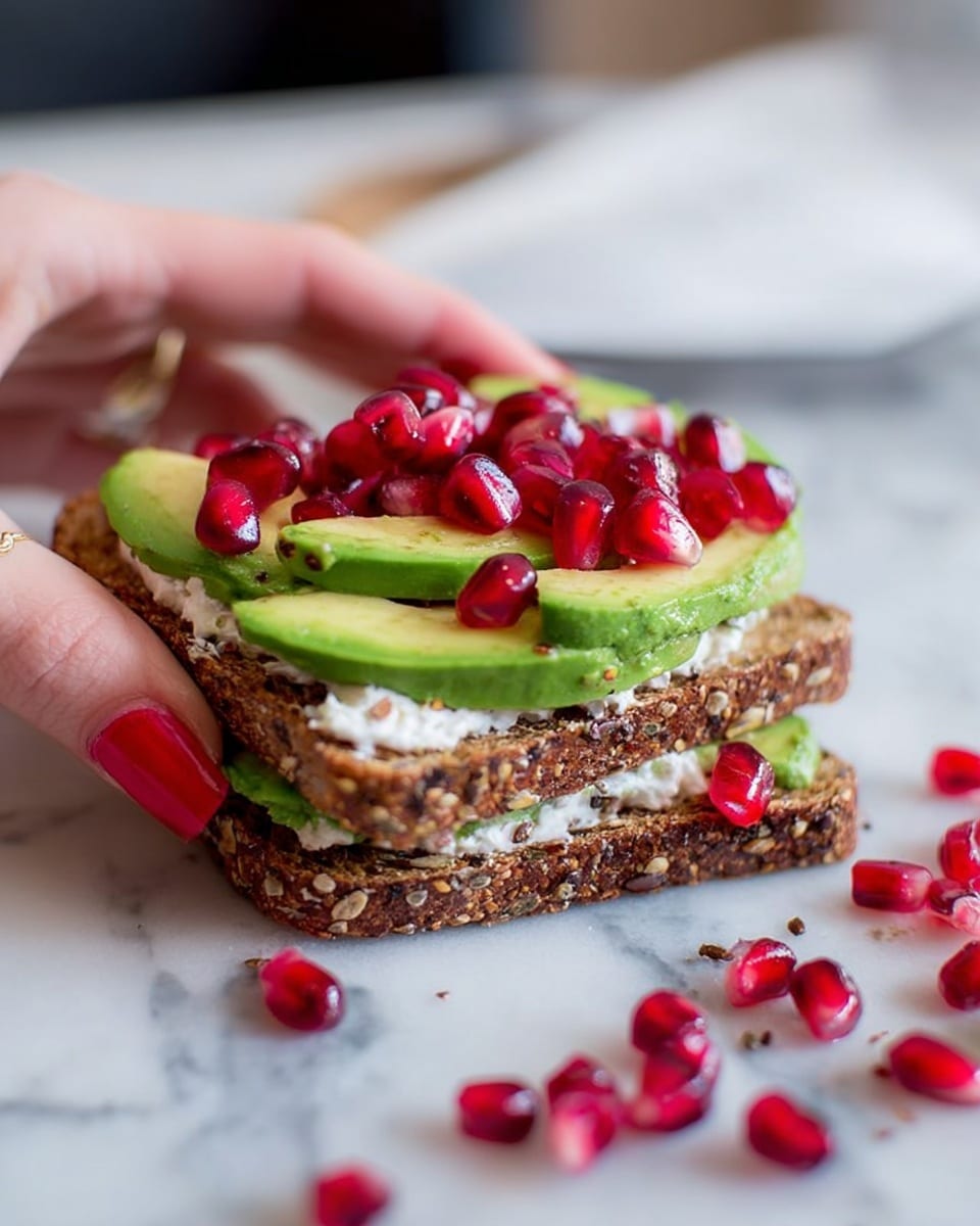 A woman's hand with red nail polish is holding a slice of seeded brown bread topped with a thin layer of white cream cheese, followed by overlapping slices of light green avocado, and sprinkled with bright red pomegranate seeds scattered on top and around the bread on a white marbled surface, with a blurred knife in the foreground. photo taken with an iphone --ar 4:5 --v 7