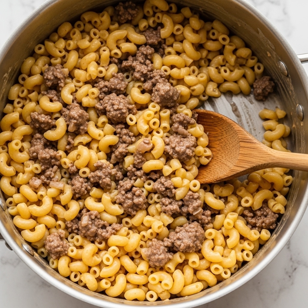 The image shows a close-up of a metal pot filled with creamy macaroni pasta mixed with ground beef. The pasta is small, curved elbow macaroni and has a smooth, slightly shiny texture from the sauce. The ground beef is cooked and crumbly, spread evenly throughout the pasta with a brownish color blending well with the light yellow pasta. A wooden spoon is partially visible stirring the mix, with its brown textured surface contrasting against the food. The whole scene is set on a white marbled texture. Photo taken with an iphone --ar 4:5 --v 7