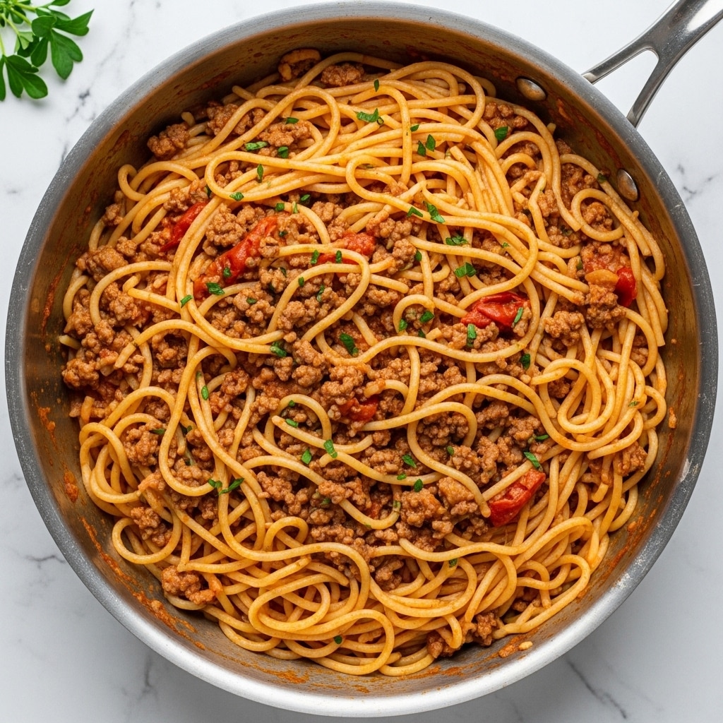 The image shows a close-up view of a large stainless steel pot filled with creamy pasta mixed with ground meat and tomato chunks. The pasta noodles are long and thick, coated in a rich orange-red sauce with bits of browned ground meat spread evenly throughout. Bright red pieces of diced tomato and small green herbs are sprinkled on top, adding pops of color and freshness. The texture looks creamy and saucy, with the noodles curling and twisting around the meat and sauce layers, all sitting inside the shiny pot against a white marbled texture. photo taken with an iphone --ar 4:5 --v 7