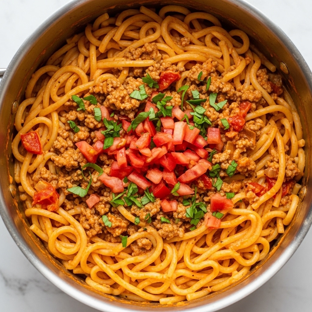 The image shows a large silver pan filled with cooked spaghetti mixed with a thick, rich meat sauce. The spaghetti noodles are long and light golden yellow, twisted and covered in a reddish-brown sauce that has chunks of ground meat and small pieces of cooked tomatoes. Small bits of green herbs are sprinkled gently across the dish, adding a touch of color. The texture of the sauce looks creamy and smooth, coating the noodles and meat evenly. The pan is placed on a white marbled surface with a small green herb sprig slightly visible at the top left corner. Photo taken with an iphone --ar 4:5 --v 7