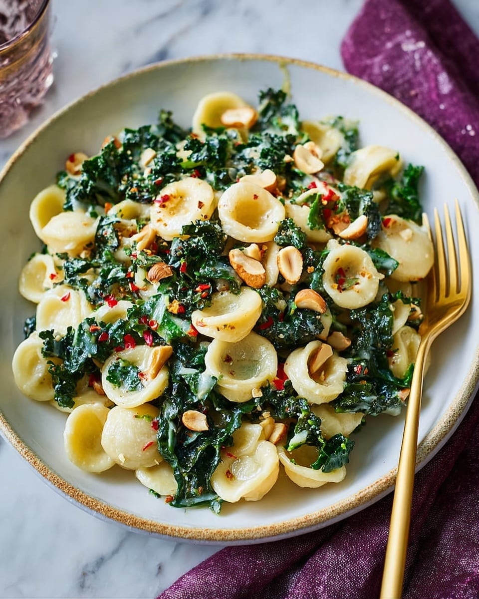 A white bowl holds a dish with two main layers: creamy light yellow orecchiette pasta that has a smooth texture, mixed generously with dark green kale leaves that look fresh and slightly wilted. Scattered throughout are small pieces of chopped nuts, light brown with a crunchy look, adding texture on top and mixed throughout. There are also tiny red chili flakes and bits of lighter green, likely herbs, spread evenly, adding color contrast. A shiny gold fork rests on the right side of the bowl, all set on a white marbled surface with a purple cloth partly visible underneath. Photo taken with an iphone --ar 4:5 --v 7