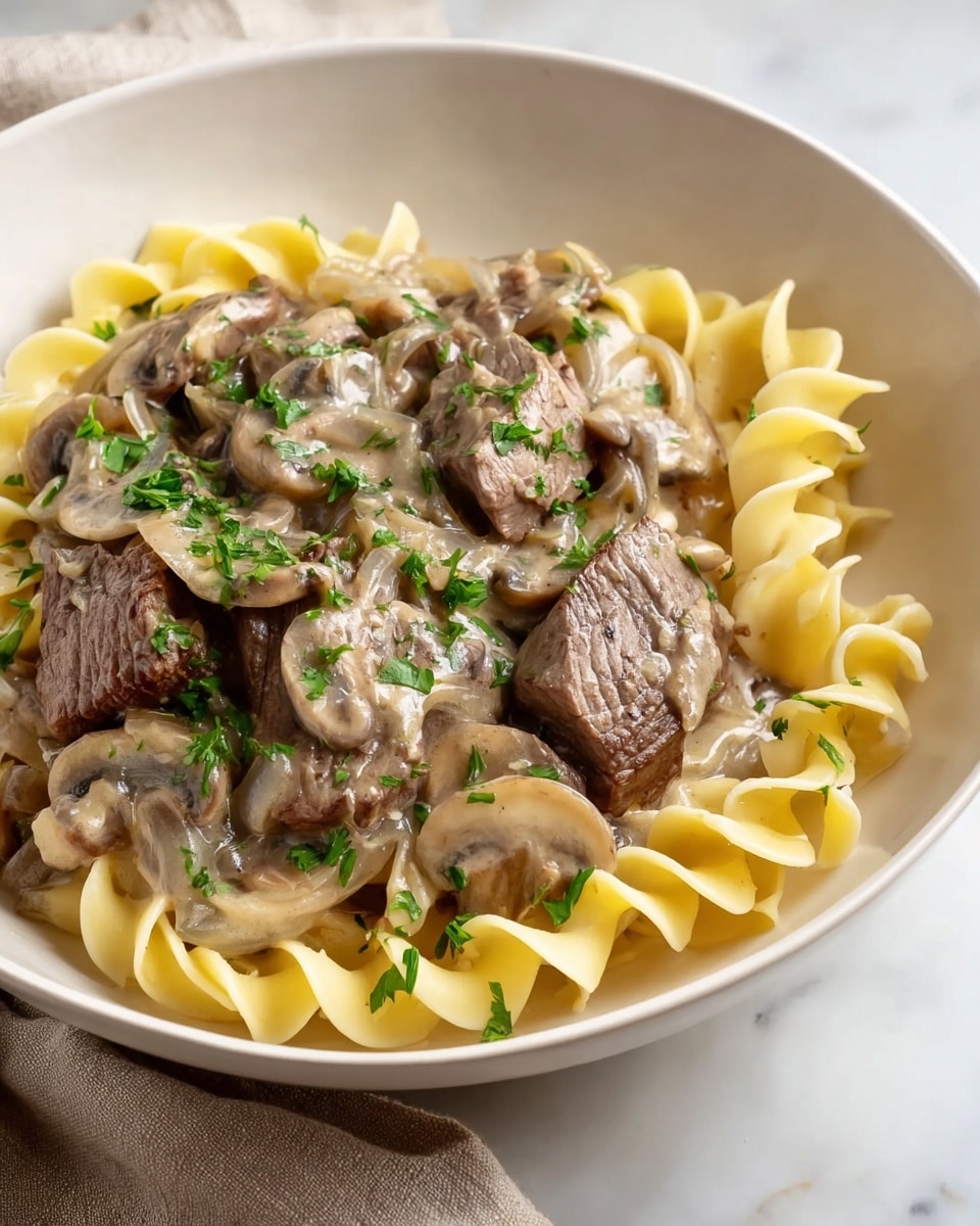 A white plate holds a serving of egg noodles at the bottom layer, pale yellow with a slightly curly and soft texture. On top of the noodles, there is a generous layer of beef stroganoff, with tender brown pieces of beef covered in a creamy, light brown sauce. Mixed into the sauce are sautéed slices of light brown mushrooms and translucent cooked onions. The dish is sprinkled with small bits of chopped green parsley, adding a fresh touch. The plate sits on a surface with a white marbled texture. photo taken with an iphone --ar 4:5 --v 7