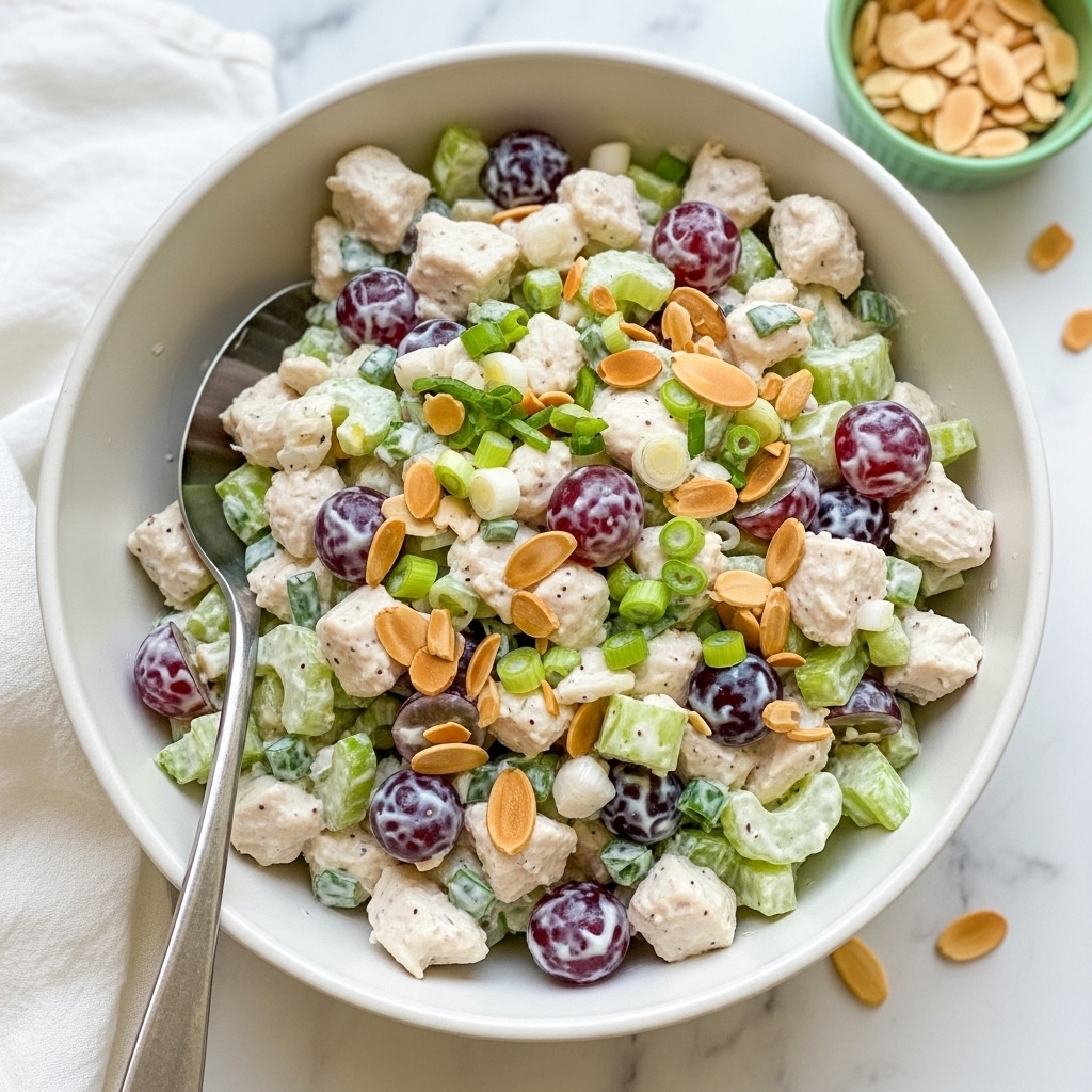 A white bowl filled with a creamy chicken salad, where chunks of cooked white chicken are mixed with bright green celery pieces and sliced red grapes, all coated in a light mayonnaise dressing. Scattered on top and throughout are thin, toasted almond slices adding a golden brown contrast. Small bits of green herbs and chopped green onions are visible, giving a fresh speckled look. A silver serving spoon is placed inside the bowl on the right side. The bowl sits on a white marbled surface with a small white cloth nearby and another small white bowl filled with toasted almond slices partially visible. Photo taken with an iphone --ar 4:5 --v 7