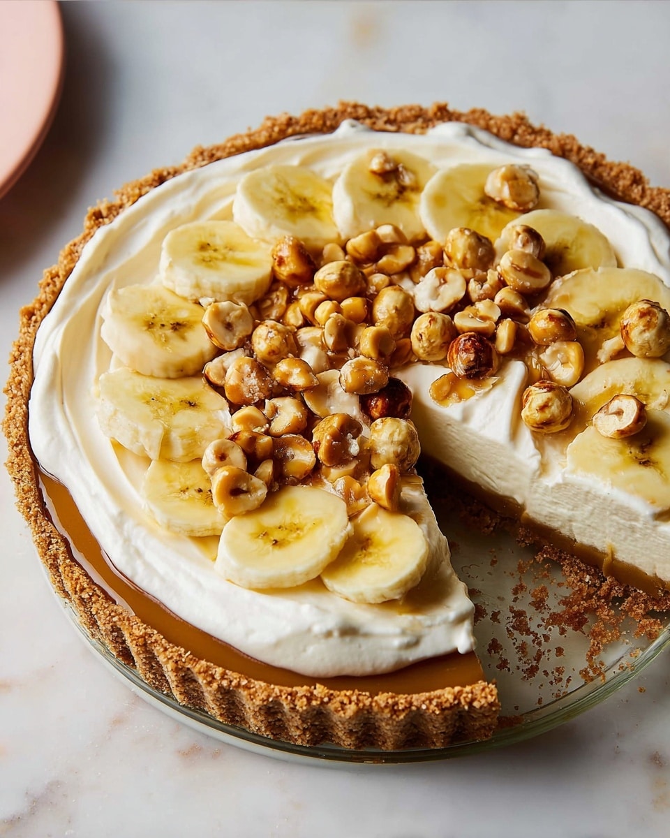 A round tart with a thick, crumbly brown crust filled with a smooth, light brown layer of caramel or toffee. On top of that is a layer of sliced bananas arranged in a circle near the edge. The center is covered with a thick swirl of creamy white whipped topping, scattered with small pieces of shiny, caramel-coated nuts. The tart sits on a white plate on a white marbled surface, with a yellow plate and a cup of coffee nearby. photo taken with an iphone --ar 4:5 --v 7