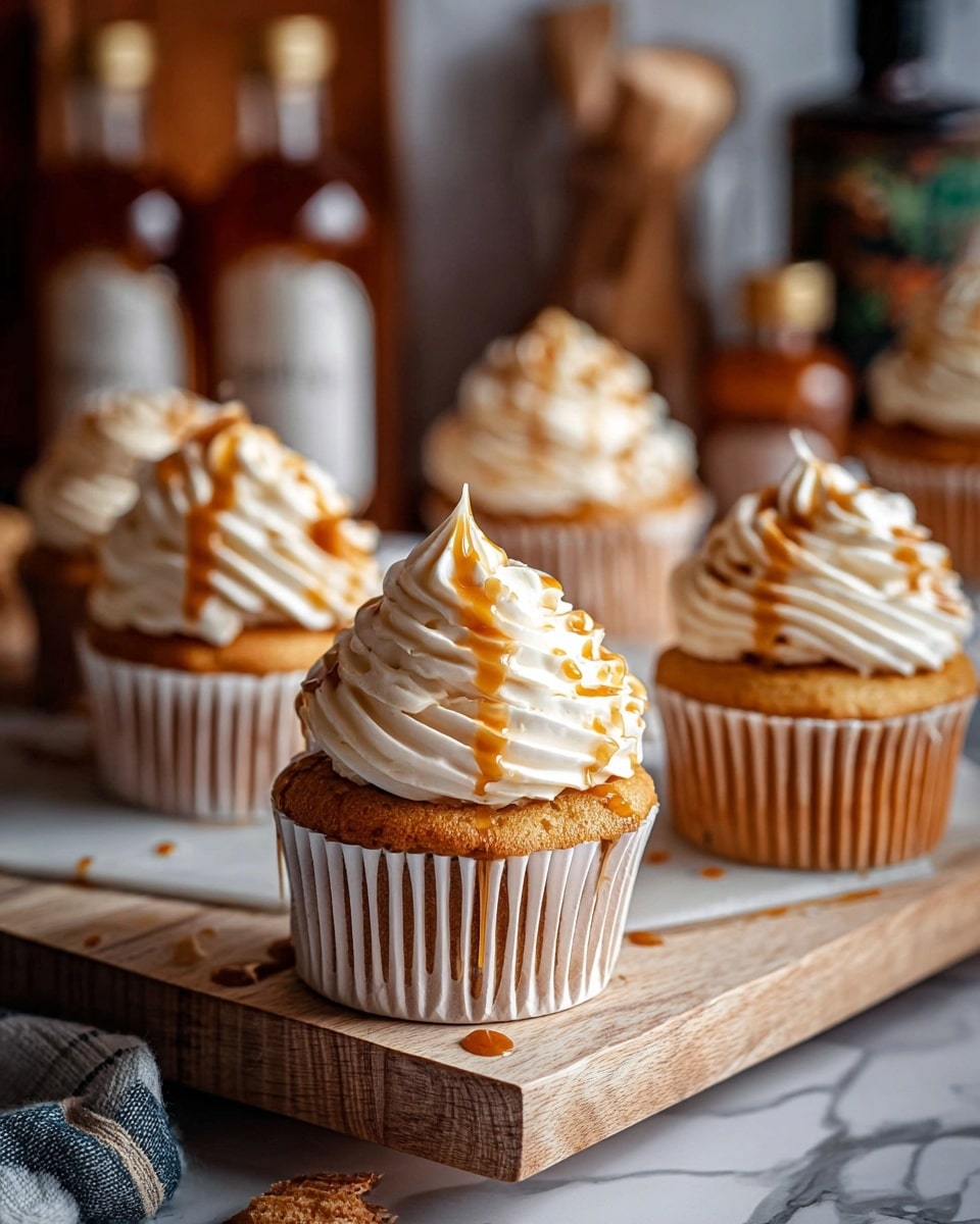 The image shows several cupcakes each with a single layer of golden brown cake in white cupcake liners. On top of each cake is a large swirl of smooth, creamy white frosting with a soft texture, drizzled with amber-brown caramel sauce that glistens. The cupcakes are arranged on a wooden board and a white stand, placed on a white marbled surface. The background has blurred bottles with dark and gold labels adding a cozy feel. Photo taken with an iphone --ar 4:5 --v 7