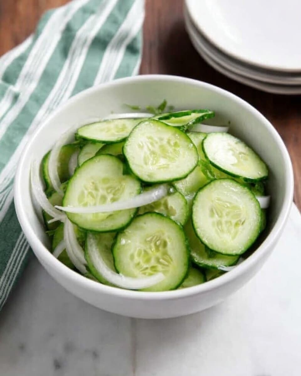 A white bowl filled with thinly sliced cucumber rounds, mixed with thin slices of white onion, sits on a wooden surface with a green-striped white napkin nearby. The cucumbers have a fresh light green color with darker green skins, and the onions add a slight translucent white contrast, creating a simple, fresh look. Photo taken with an iphone --ar 4:5 --v 7