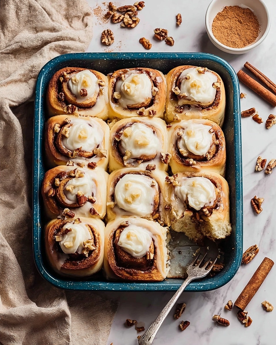 A close-up of a blue baking dish filled with twelve soft cinnamon rolls arranged in a 3 by 4 grid, each roll showing three visible layers: a pale golden-brown outer dough, a dark cinnamon sugar swirl inside, and a creamy white dollop of icing on top sprinkled with chopped pecans. One cinnamon roll in the lower right corner is partly eaten, showing the fluffy, light texture inside, and a fork is resting in the empty space. Scattered around the dish on a white marbled surface are pecan pieces, cinnamon sticks, and a small bowl filled with ground cinnamon, with a beige linen cloth draped loosely on the left side. photo taken with an iphone --ar 4:5 --v 7