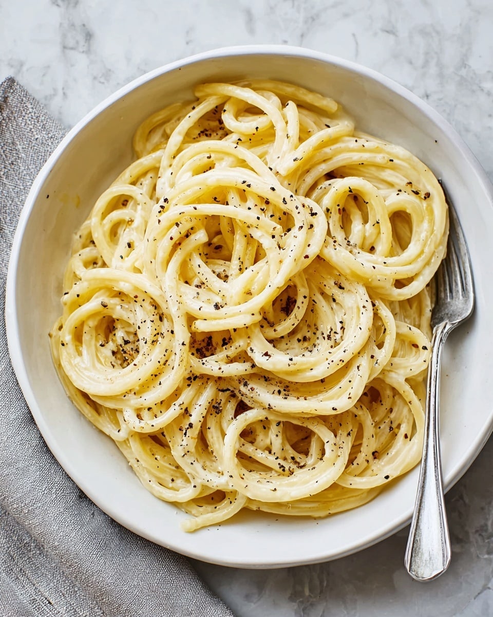 A white bowl filled with thick, smooth spaghetti noodles coated in a light, slightly shiny creamy sauce. The noodles are arranged in loose, round piles with a few strands overlapping. The pasta is topped with a sprinkle of coarse black pepper, adding specks of dark contrast over the pale yellow noodles. The bowl is placed on a white marbled surface with a soft, light gray cloth and a silver fork resting beside it. photo taken with an iphone --ar 4:5 --v 7