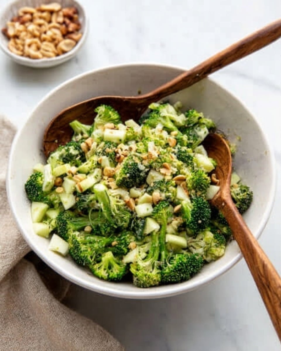 A white bowl on a white marbled surface holds a broccoli salad with small broccoli pieces mixed with bits of almonds and other small chopped nuts, giving a mix of green and light brown colors. Two wooden salad spoons rest inside the bowl, angled outwards. In the background, a small white bowl with chopped nuts is blurred, along with a beige napkin. The overall look is fresh and natural. Photo taken with an iphone --ar 4:5 --v 7